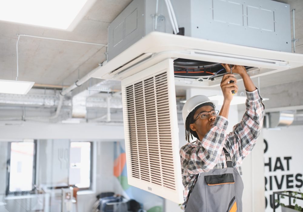 Technician in a hard hat and uniform working on an air conditioning unit in a modern office, ensuring proper ventilation and cooling